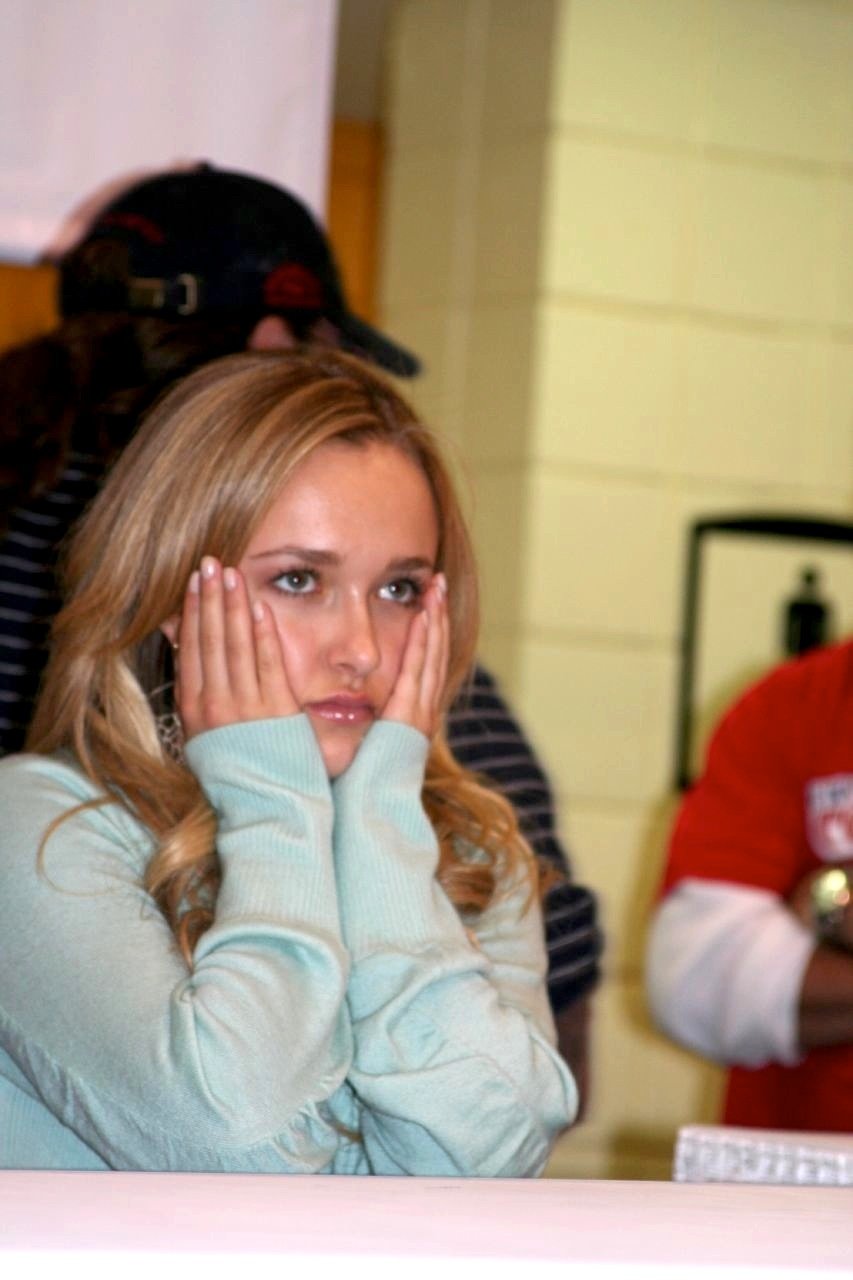 Young woman with long blonde hair and a pale blue sweater sits at a table indoors, cupping her cheeks with both hands and looking off-camera; blurred people and cream-colored tiled walls are visible in the background.