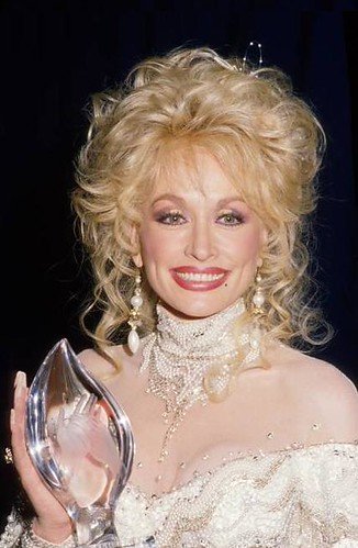 Blonde woman wearing statement jewelry and a lacy top, smiling and holding a clear glass award.