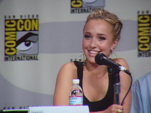 Blonde woman with hair in an updo wearing a black sleeveless top, smiling into a tabletop microphone. A small water bottle and a panel nameplate sit on the table in front of her; a Comic-Con logo backdrop is visible behind her.