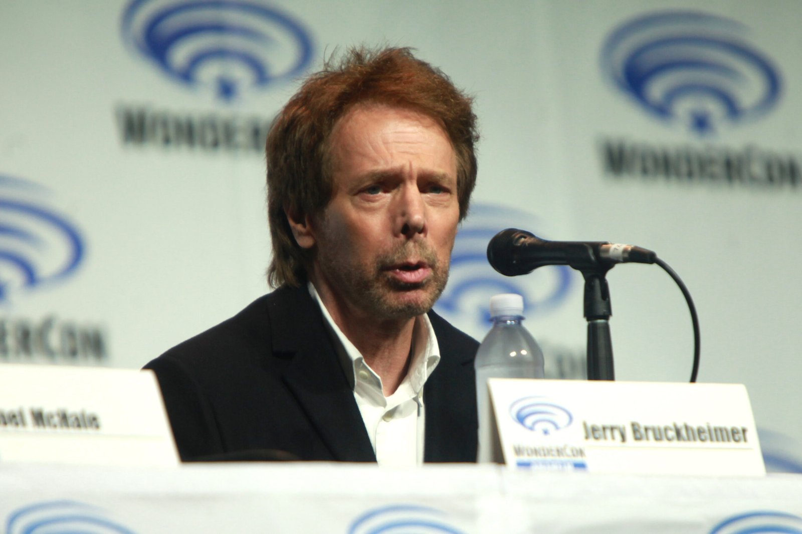 Man identified as Jerry Bruckheimer speaking into a microphone at a panel table with a WonderCon logo backdrop; a name placard and water bottle sit on the table.