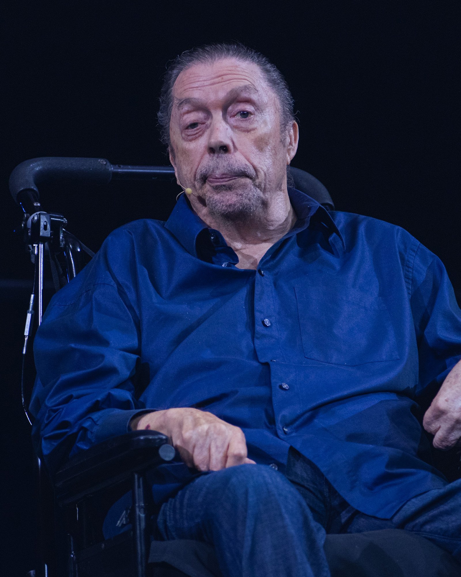 An older man seated in a wheelchair onstage wearing a button-up shirt and a small headset microphone.