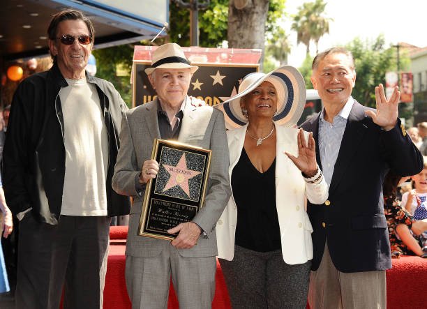 Four people standing side by side at a Walk of Fame ceremony; one person holds a plaque with a star while the others stand close and wave.